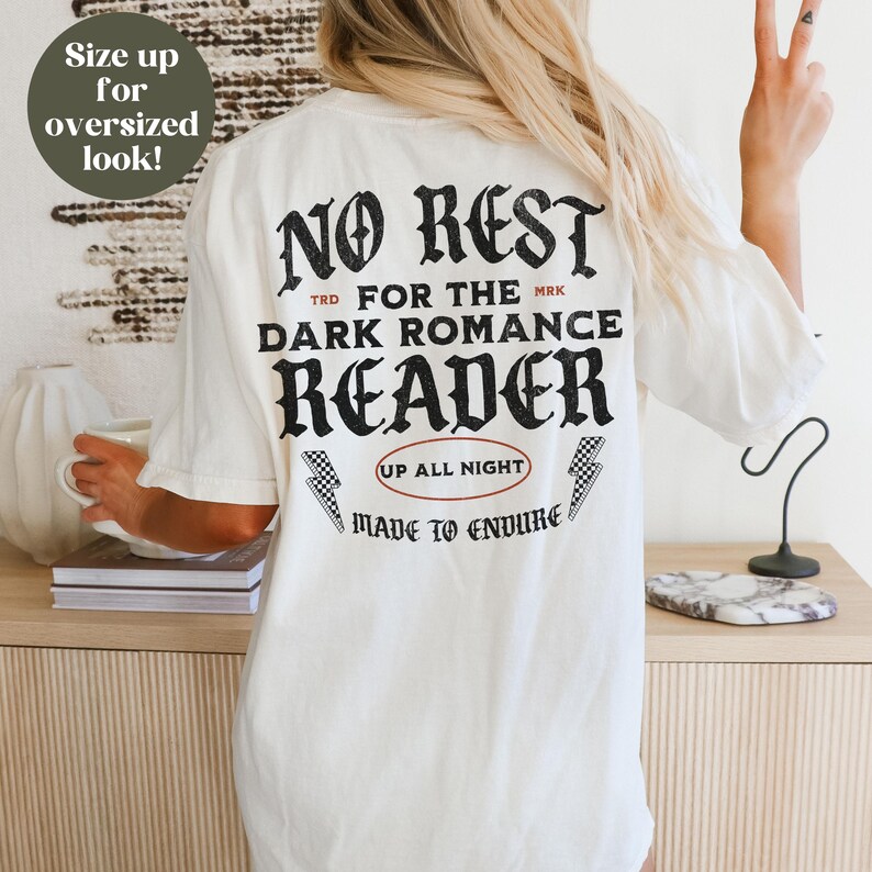 May include: White t-shirt with black text that reads "NO REST FOR THE DARK ROMANCE READER" with a red circle around the text "UP ALL NIGHT" and black and white lightning bolts on either side. The text "MADE TO ENDURE" is below the circle.
