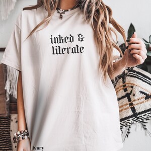 May include: Ivory-colored t-shirt with the text "inked & literate" in a gothic font. The shirt has short sleeves and a relaxed fit. The image shows the shirt on a person.