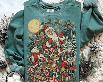 Puede incluir: Una sudadera de cuello redondo en color verde claro con un estampado gráfico festivo de Papá Noel, un muñeco de nieve, un reno y un cascanueces. El diseño incluye una luna llena, renos voladores, un trineo de madera y un pueblo.