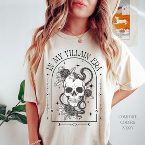 May include: A cream-colored t-shirt with a black and white graphic of a skull with a snake wrapped around it. The graphic is surrounded by flowers and stars. The text "In My Villain Era" is written in a black arch above the graphic. The text "Comfort Colors Ivory" is written in black in the bottom right corner of the image.