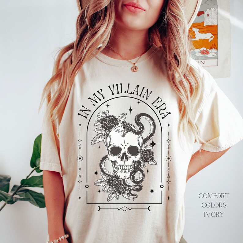 May include: A cream-colored t-shirt with a black and white graphic of a skull with a snake wrapped around it. The graphic is surrounded by flowers and stars. The text "In My Villain Era" is written in a black arch above the graphic. The text "Comfort Colors Ivory" is written in black in the bottom right corner of the image.