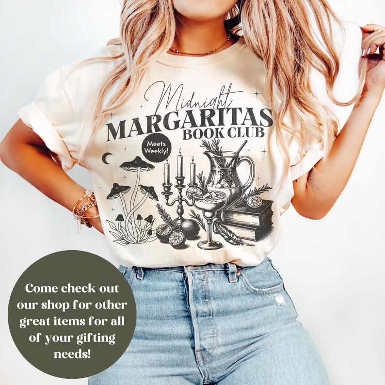 Puede incluir: Camiseta color crema con un dise&ntilde;o gr&aacute;fico en blanco y negro. El dise&ntilde;o presenta el texto "Midnight Margaritas Book Club" con ilustraciones de setas, velas, una jarra y un c&oacute;ctel. La camiseta tambi&eacute;n tiene el texto "Meets Weekly!"