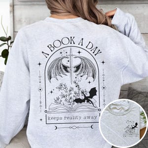 May include: Gray sweatshirt with a black and white graphic design featuring a book, a dragon, wings, and stars. The text reads "A Book A Day Keeps Reality Away".