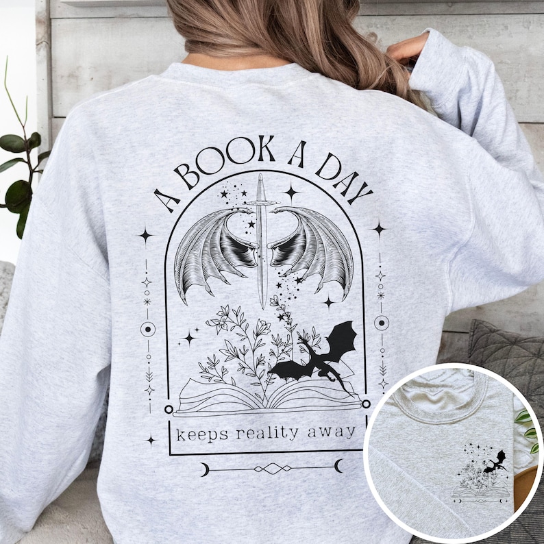 May include: Gray sweatshirt with a black and white graphic design featuring a book, a dragon, wings, and stars. The text reads "A Book A Day Keeps Reality Away".