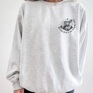 May include: A light gray sweatshirt with a crew neck. The sweatshirt features a small graphic on the upper left chest. The graphic is a black and white illustration of an open book with a skeleton hand and the text "Morally Grey is my Favorite Color."