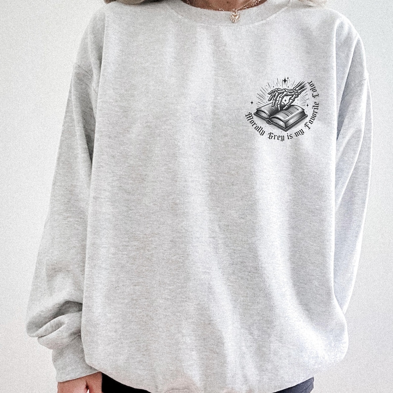 May include: A light gray sweatshirt with a crew neck. The sweatshirt features a small graphic on the upper left chest. The graphic is a black and white illustration of an open book with a skeleton hand and the text "Morally Grey is my Favorite Color."