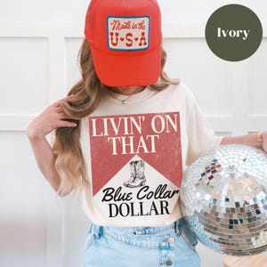 May include: A white t-shirt with a red and white graphic that says "Livin' on That Blue Collar Dollar". The graphic features a pair of cowboy boots. The model is wearing a red trucker hat with a white "Made in the U.S.A." patch.