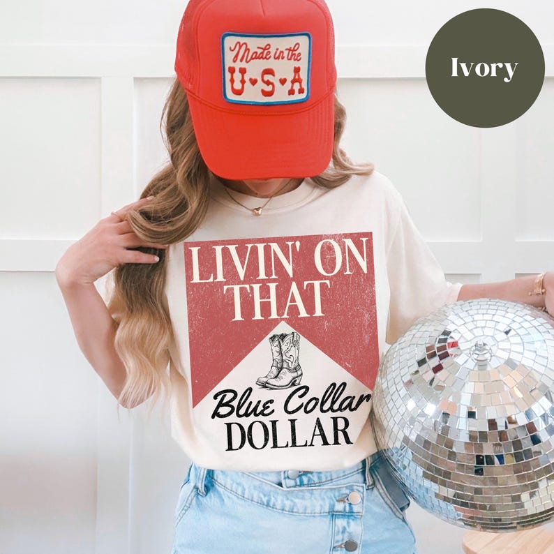 May include: A white t-shirt with a red and white graphic that says "Livin' on That Blue Collar Dollar". The graphic features a pair of cowboy boots. The model is wearing a red trucker hat with a white "Made in the U.S.A." patch.