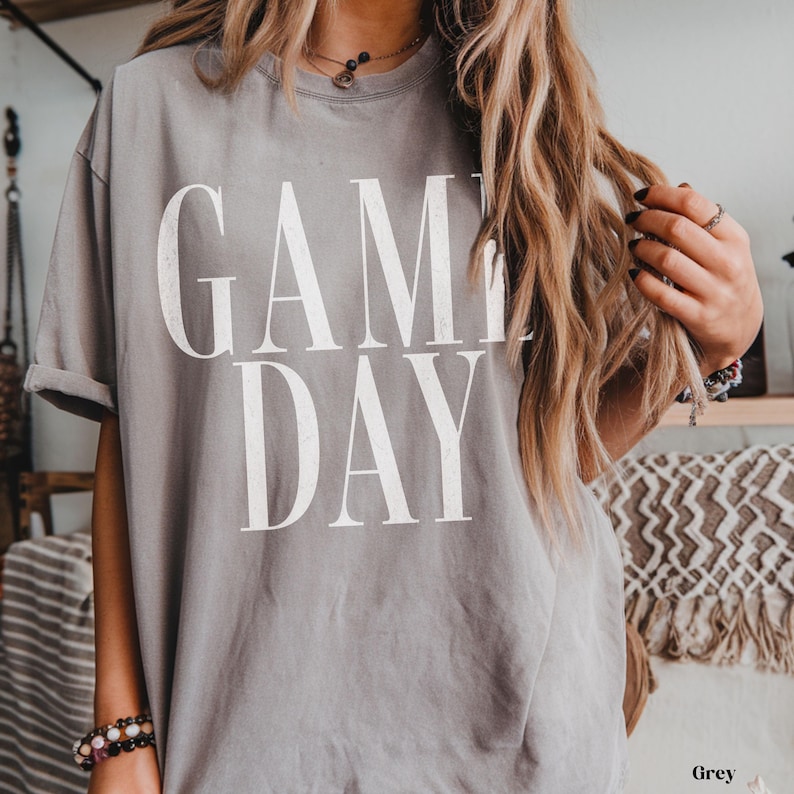 May include: A grey short-sleeved t-shirt featuring the text "GAME DAY" printed in large, white, distressed-style capital letters across the chest. The shirt has a crew neckline and a relaxed fit, with the fabric showing a soft, worn-in texture.
