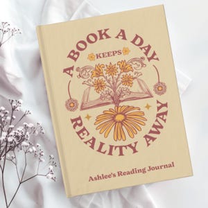 Puede incluir: Un diario de lectura beige con el texto "A Book A Day Keeps Reality Away" en un dise&ntilde;o circular. El dise&ntilde;o incluye flores y una ilustraci&oacute;n de libro abierto. El diario tambi&eacute;n tiene el texto "Ashlee's Reading Journal" en la parte inferior.