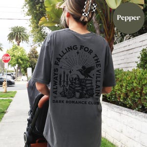 May include: A dark grey t-shirt with a black graphic print of a skull, raven, and candles. The text "Always Falling For The Villain" and "Dark Romance Club" are printed on the shirt.