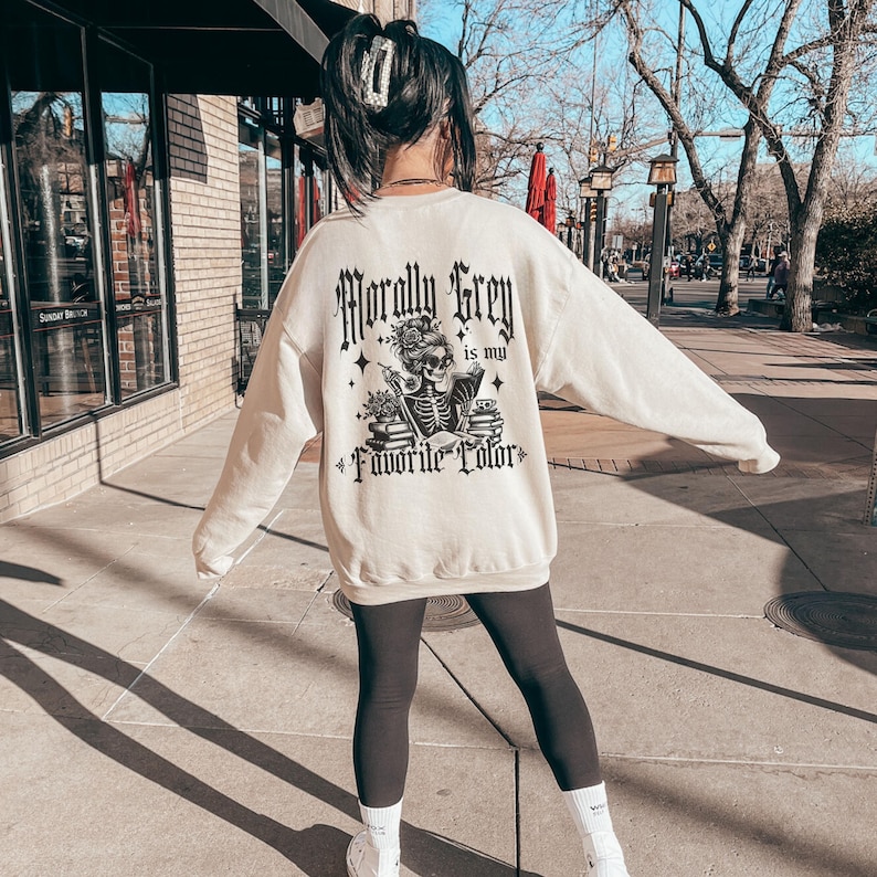 May include: Cream-colored sweatshirt with a graphic on the back that reads "Morally Grey is my Favorite Color." The graphic features a skeleton with a floral headpiece, surrounded by stars and books. The sweatshirt is paired with black leggings.