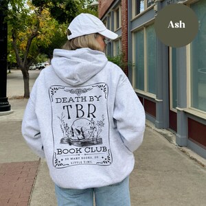 K&ouml;nnte beinhalten: Ein grauer Kapuzenpullover mit einem schwarzen grafischen Aufdruck auf der R&uuml;ckseite. Der Aufdruck zeigt einen Sch&auml;del, der ein Buch liest, mit dem Text "Death by TBR Book Club. So many books, so little time."
