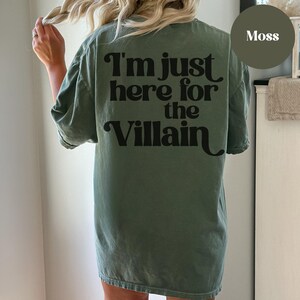 Puede incluir: Una camiseta verde musgo con el texto "I'm just here for the Villain" impreso en negro en la parte posterior.