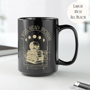 K&ouml;nnte beinhalten: Eine schwarze Keramiktasse mit einem wei&szlig;en Design, das einen Sch&auml;del zeigt, der auf einem Stapel B&uuml;cher sitzt, mit dem Text "A well read woman is a dangerous creature" und einem Halbmond-Design.