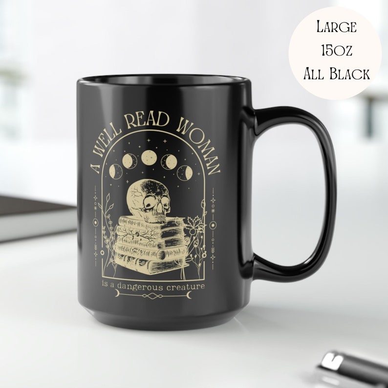 K&ouml;nnte beinhalten: Eine schwarze Keramiktasse mit einem wei&szlig;en Design, das einen Sch&auml;del zeigt, der auf einem Stapel B&uuml;cher sitzt, mit dem Text "A well read woman is a dangerous creature" und einem Halbmond-Design.