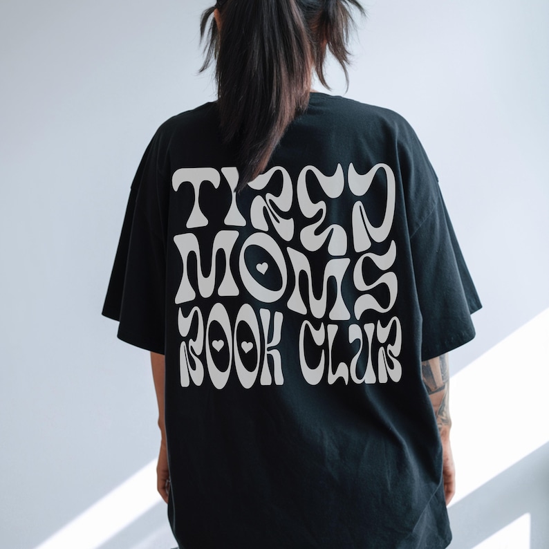A woman turned around wearing a black shirt that reads "TIRED MOMS BOOK CLUB" in a white psychedelic-like font.
