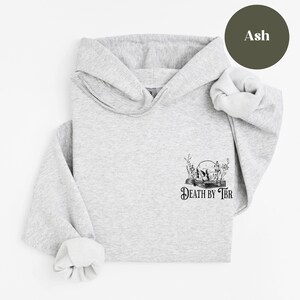 K&ouml;nnte beinhalten: Ein grauer Kapuzenpullover mit einer schwarz-wei&szlig;en Grafik eines Sch&auml;dels auf einem Buch mit dem Text "Death by TBR" darunter. Der Pullover befindet sich auf einem wei&szlig;en Hintergrund.