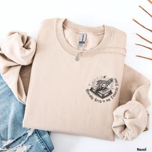 May include: A sand-colored sweatshirt with a crew neck and long sleeves. The graphic on the front features a skeleton hand reaching for an open book, with the text "Morally Grey is my Favorite Color". A silver necklace with a sun pendant is also visible.