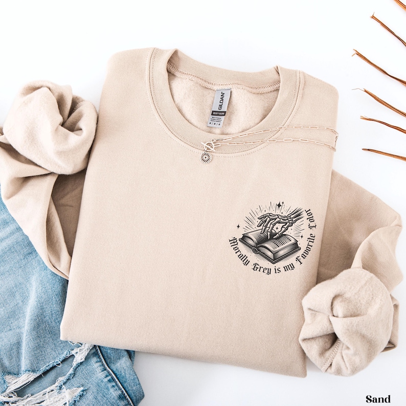 May include: A sand-colored sweatshirt with a crew neck and long sleeves. The graphic on the front features a skeleton hand reaching for an open book, with the text "Morally Grey is my Favorite Color". A silver necklace with a sun pendant is also visible.