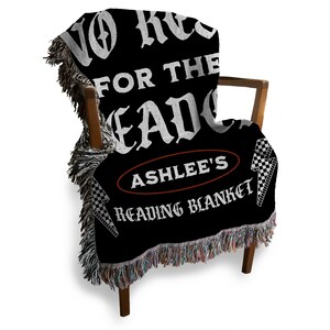 May include: Black throw blanket draped over a wooden chair. The blanket features white text that reads "NO REGRETS FOR THE DEAD" and "ASHLEE'S READING BLANKET" in a decorative font. The blanket has a fringed edge.