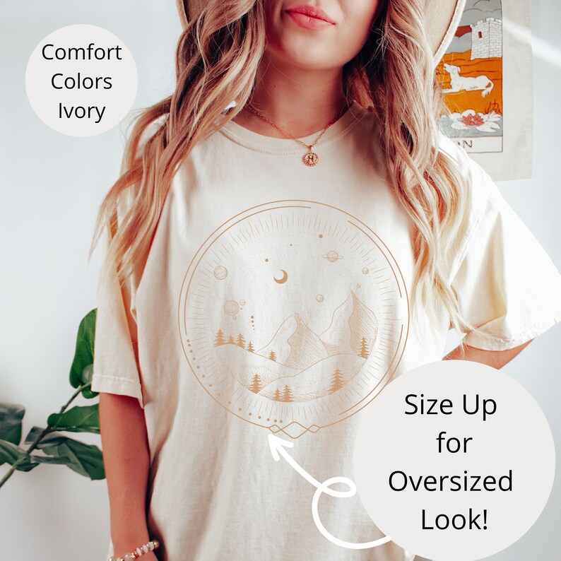 Puede incluir: Una camiseta blanca con un gr&aacute;fico de una cordillera, luna y estrellas dentro de un c&iacute;rculo. El texto "Size Up for Oversized Look!" est&aacute; en la esquina inferior derecha de la imagen.