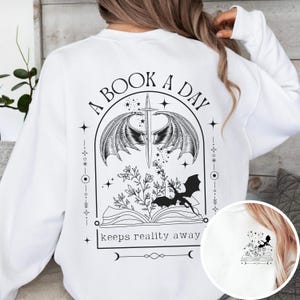 May include: White sweatshirt with a black graphic print of a book with wings, a sword, and a dragon. The text "A Book A Day Keeps Reality Away" is printed below the graphic.