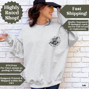 May include: Ash-colored sweatshirt with a crew neck. The graphic on the front features a book with a skeleton hand and the text "Morally Grey is my Favorite Color". The sweatshirt has long sleeves and is made of a soft material.