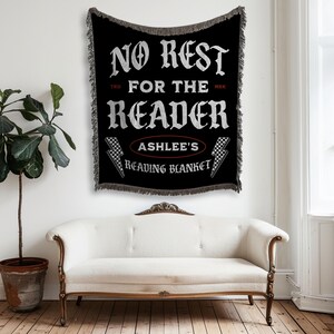 May include: Black woven throw blanket with white text that reads "NO REST FOR THE READER" and "ASHLEE'S READING BLANKET". The blanket has a fringed edge and lightning bolt graphics. The blanket is hanging on a white wall above a white couch.