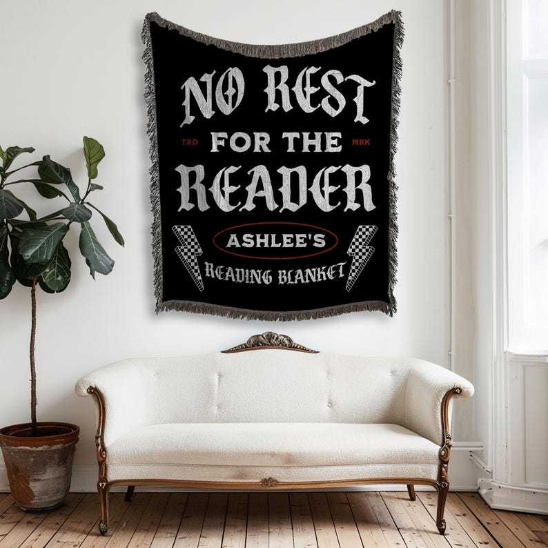 May include: Black woven throw blanket with white text that reads "NO REST FOR THE READER" and "ASHLEE'S READING BLANKET". The blanket has a fringed edge and lightning bolt graphics. The blanket is hanging on a white wall above a white couch.