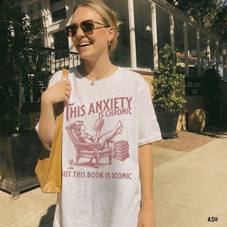 K&ouml;nnte beinhalten: Ein wei&szlig;es T-Shirt mit einer roten Grafik einer Frau, die in einem Sessel ein Buch liest. Der Text auf dem Shirt lautet "This anxiety is chronic but this book is iconic."