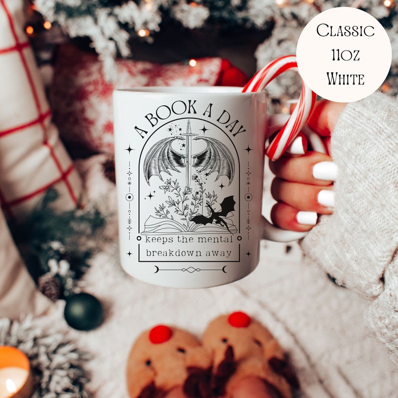 May include: White ceramic mug with a black and white illustration of a book, sword, and dragon. The text reads "A Book A Day keeps the mental breakdown away".