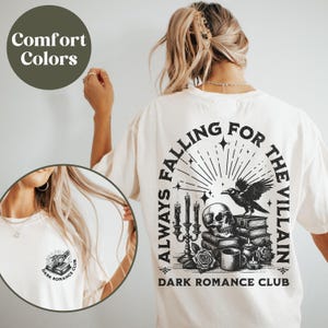 May include: White t-shirt with a black graphic print on the back that says "Always Falling for the Villain" with a skull, raven, and candles. The front of the shirt has a small black graphic print of a book with the text "Dark Romance Club".