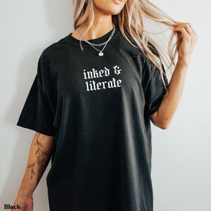 May include: Black t-shirt with the text "inked & literate" in a gothic font. The shirt has short sleeves and a relaxed fit. The model is wearing a silver necklace.