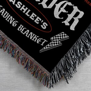 May include: Black woven blanket with white text that reads "ASHLEE'S READING BLANKET" and "FOREVER". A checkered lightning bolt graphic is also visible. The blanket has a fringed edge with multiple colors.