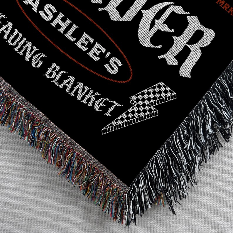 May include: Black woven blanket with white text that reads "ASHLEE'S READING BLANKET" and "FOREVER". A checkered lightning bolt graphic is also visible. The blanket has a fringed edge with multiple colors.