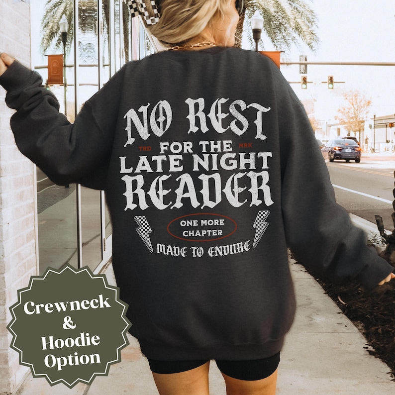 Peut inclure: Sweat-shirt gris fonc&eacute; avec l'inscription blanche "NO REST FOR THE LATE NIGHT READER". Le design comprend des &eacute;clairs et la phrase "ONE MORE CHAPTER MADE TO ENDURE". Une &eacute;tiquette verte indique les options col rond et sweat &agrave; capuche.