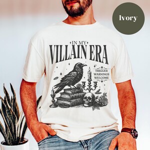May include: A white t-shirt with a black and white graphic design featuring a raven, books, and a skull. The text on the shirt reads "In My Villain Era" and "Trigger Warnings Welcome".