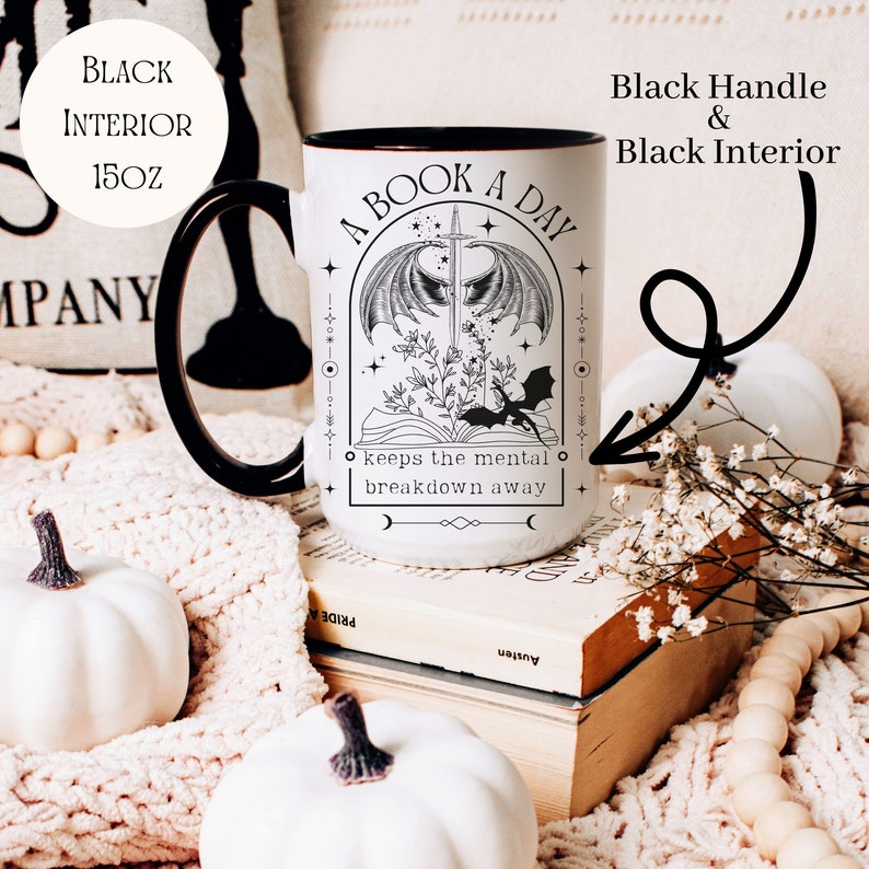 May include: White ceramic mug with a black interior and handle. The mug features a black and white illustration of a book with a dragon and a sword. The text on the mug reads "A Book A Day Keeps The Mental Breakdown Away".