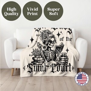 May include: A white throw blanket with a black and white illustration of a skeleton reading a book. The text "of the Smut Court" is printed on the blanket. The blanket is made in the USA.