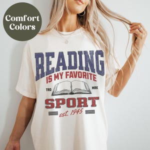 May include: A white t-shirt with a vintage-style graphic that says "Reading is my favorite sport est. 1945". The graphic features a blue and red color scheme with a stylized open book.
