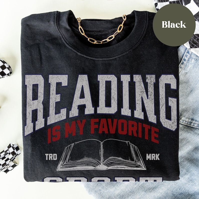 May include: Black t-shirt with a vintage-style graphic that says "Reading is my favorite sport". The graphic features a book with the words "TRD" and "MRK" on either side.