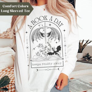 May include: White long-sleeved t-shirt with a black graphic design featuring a book, a dragon, wings, a sword, and the text "A BOOK A DAY keeps reality away".