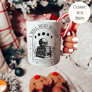 K&ouml;nnte beinhalten: Wei&szlig;e Keramiktasse mit einem schwarz-wei&szlig;en Design. Das Design zeigt einen Sch&auml;del mit einem Stapel B&uuml;cher, einen zunehmenden Mond und den Text "A well read woman is a dangerous creature".