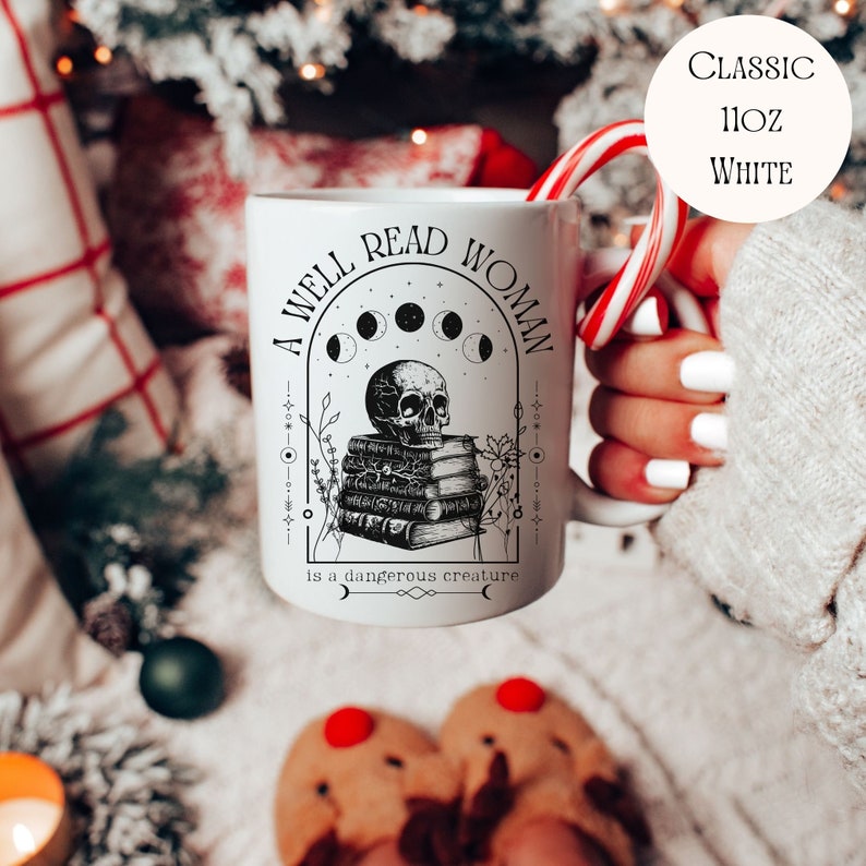 K&ouml;nnte beinhalten: Wei&szlig;e Keramiktasse mit einem schwarz-wei&szlig;en Design. Das Design zeigt einen Sch&auml;del mit einem Stapel B&uuml;cher, einen zunehmenden Mond und den Text "A well read woman is a dangerous creature".