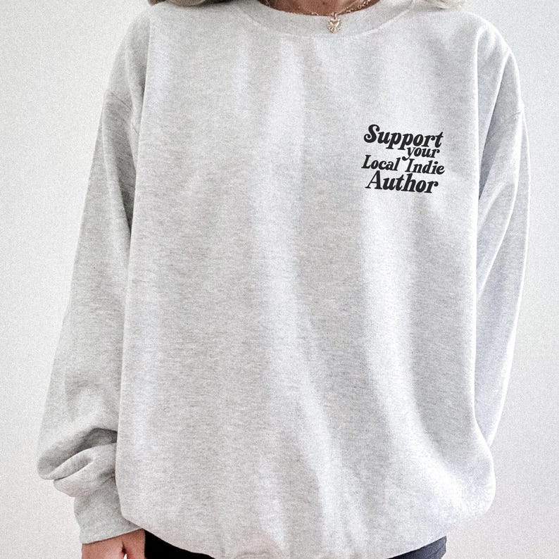May include: A light gray sweatshirt with long sleeves. The text "Support your Local Indie Author" is printed in black on the upper right chest area. The sweatshirt is made of a soft material.