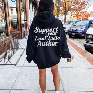 May include: Black hoodie with the text "Support your Local Indie Author" in white script. The hoodie is worn by a person walking on a sidewalk. The background includes a street with parked cars and autumn foliage.