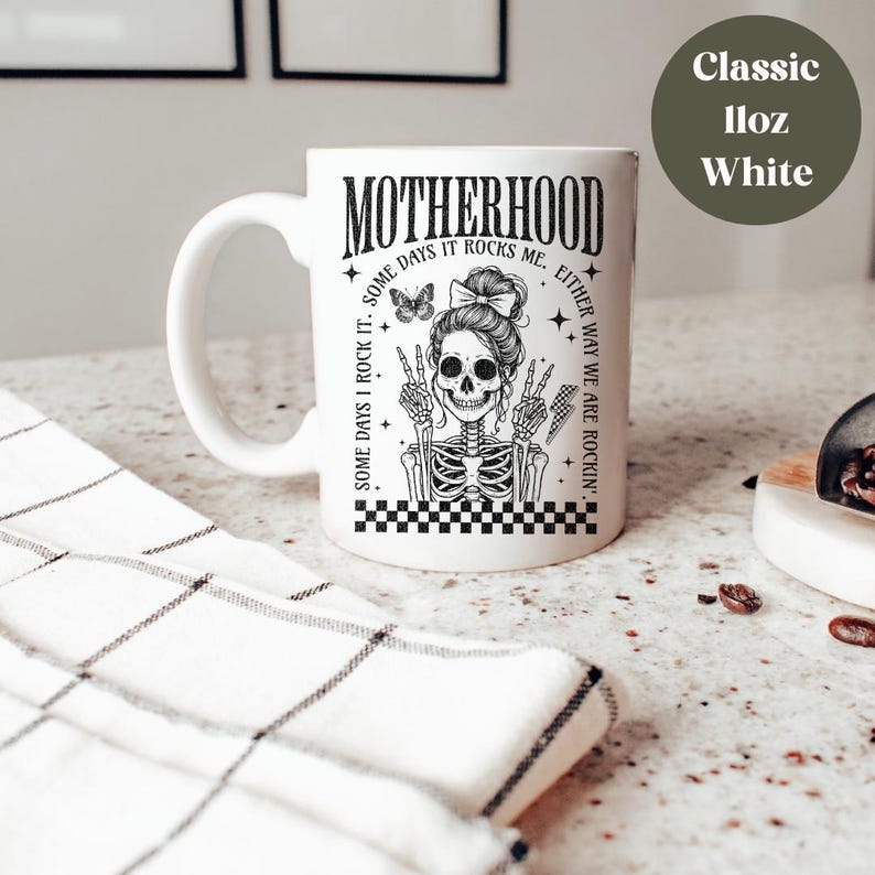 May include: White ceramic mug with a black and white illustration of a skeleton with a bow in its hair. The text "MOTHERHOOD" is printed in black on the mug. The text "SOME DAYS I ROCK IT. SOME DAYS IT ROCKS ME. EITHER WAY WE ARE ROCKIN'" is printed in black around the skeleton.