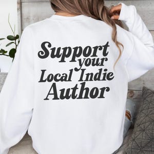 May include: White sweatshirt with the text "Support your Local Indie Author" in black, bold lettering. The sweatshirt has long sleeves and a crew neck. The image shows the back of the sweatshirt.