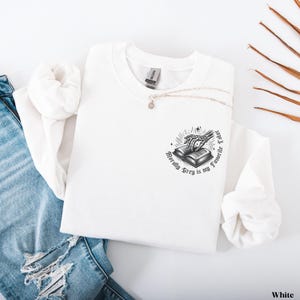 May include: White sweatshirt with a crew neck and long sleeves. The graphic on the front features a skeleton hand reaching for an open book, with the text "Morally Grey is my Favorite Color" in a circular design. A silver necklace is draped across the sweatshirt.